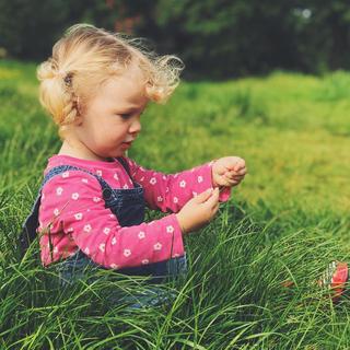 A little girl sat in the gradd wearing a bright pink flowery t-shirt and dungarees