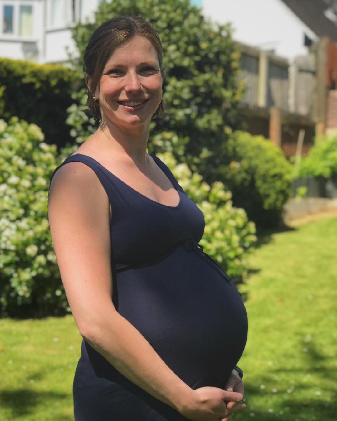 A pregnant woman standing holding her belly in a garden. 