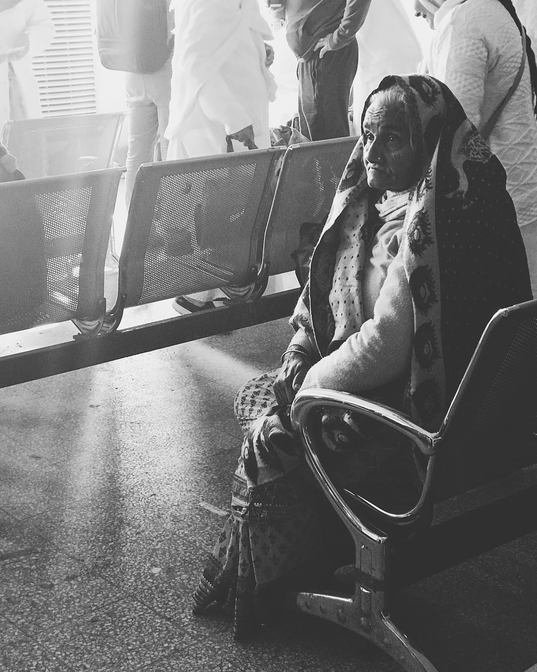 An old woman sitting in an airport chair in black and white