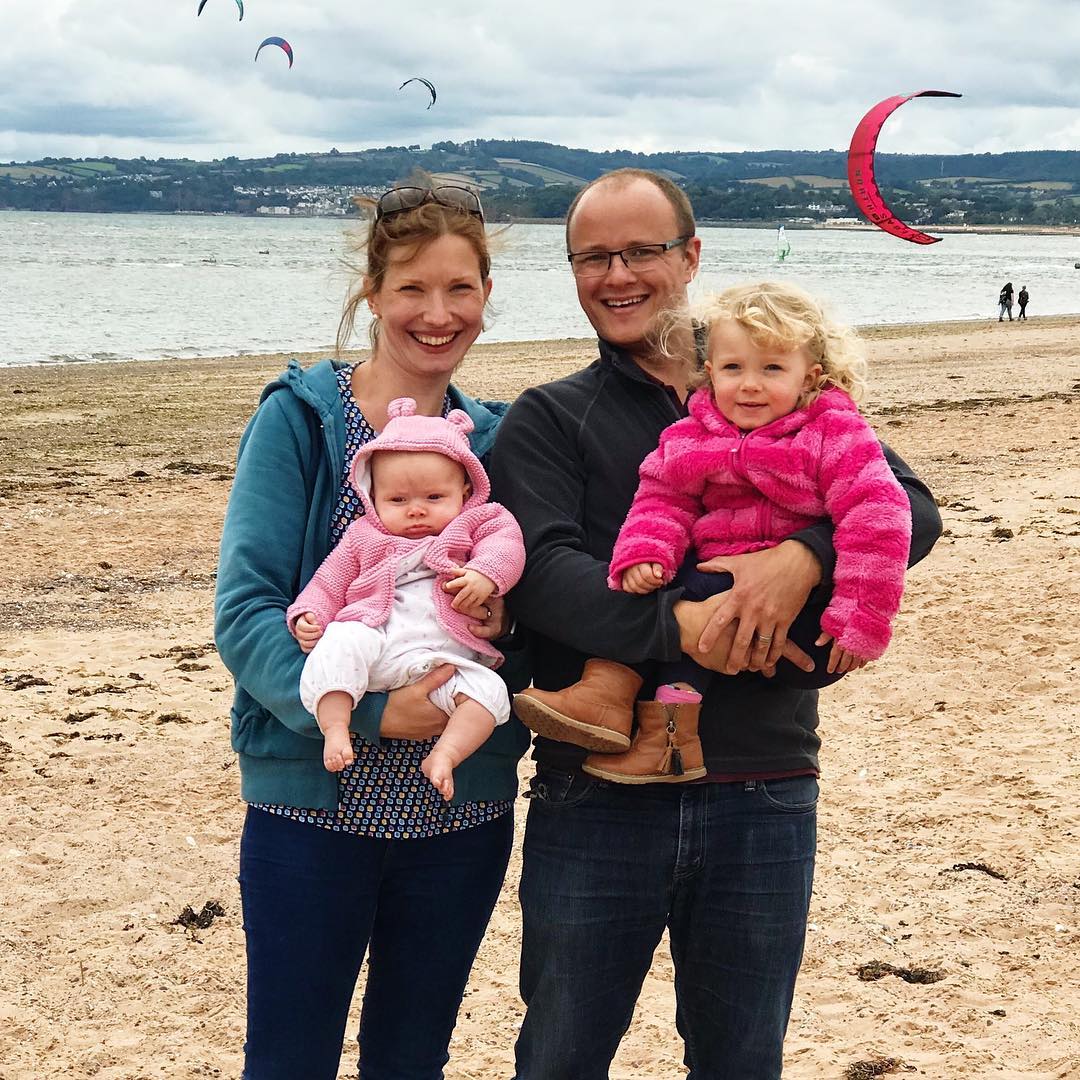 A family photo of a husband and wife holding two girls with the sea in the background.