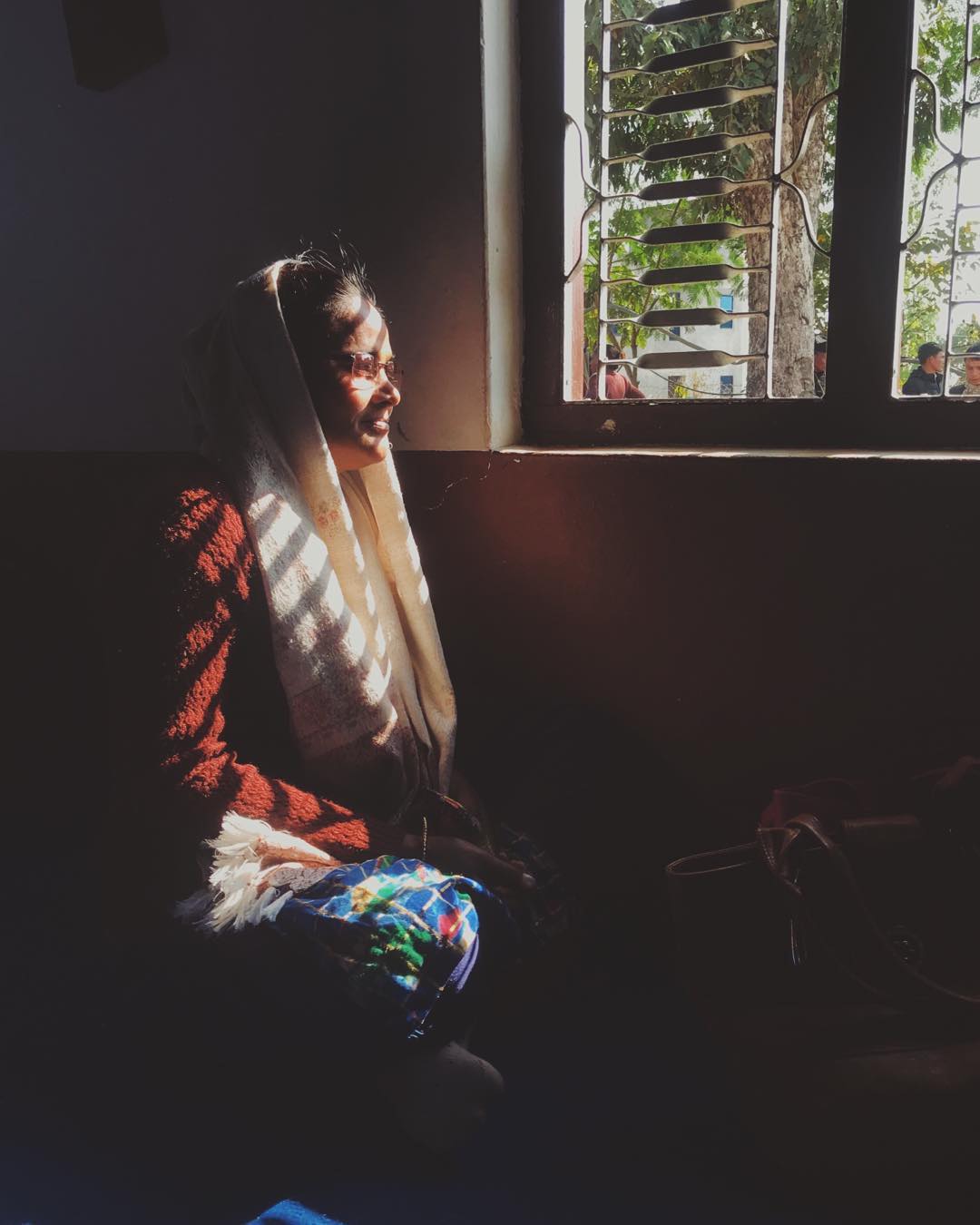 A Nepalese lady sits on the floor next to a window, the morning light casts shadows across her face.