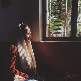 A Nepalese lady sits on the floor next to a window, the morning light casts shadows across her face.