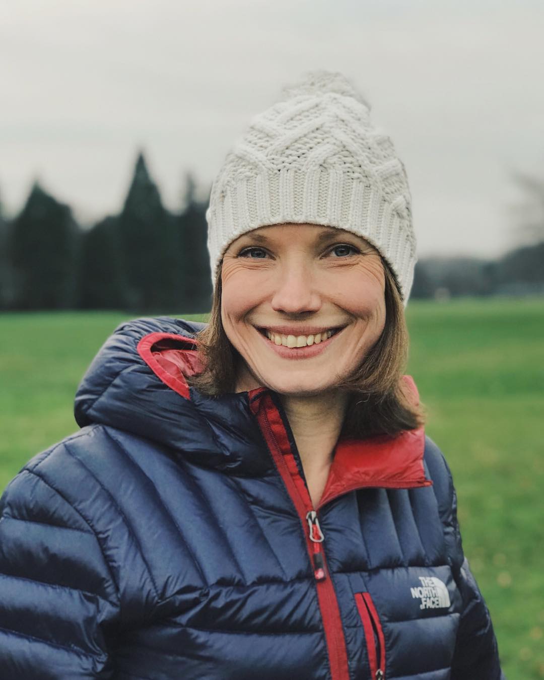 A woman smiling wearing a white bobble hat and a blue and red down jacket