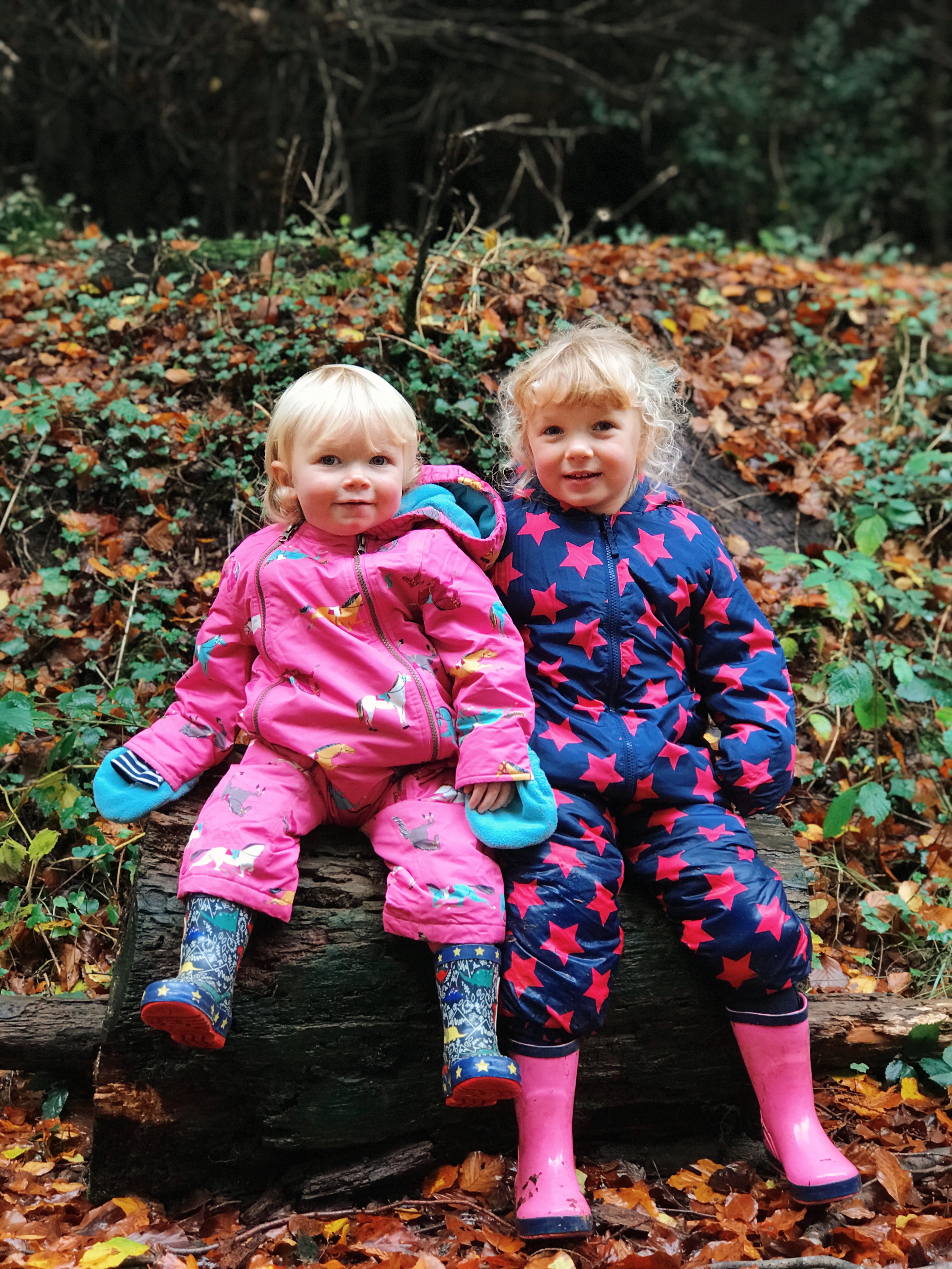Two girls sat on a log in colourful all in ones and wellies.