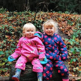 Two girls sat on a log in colourful all in ones and wellies.