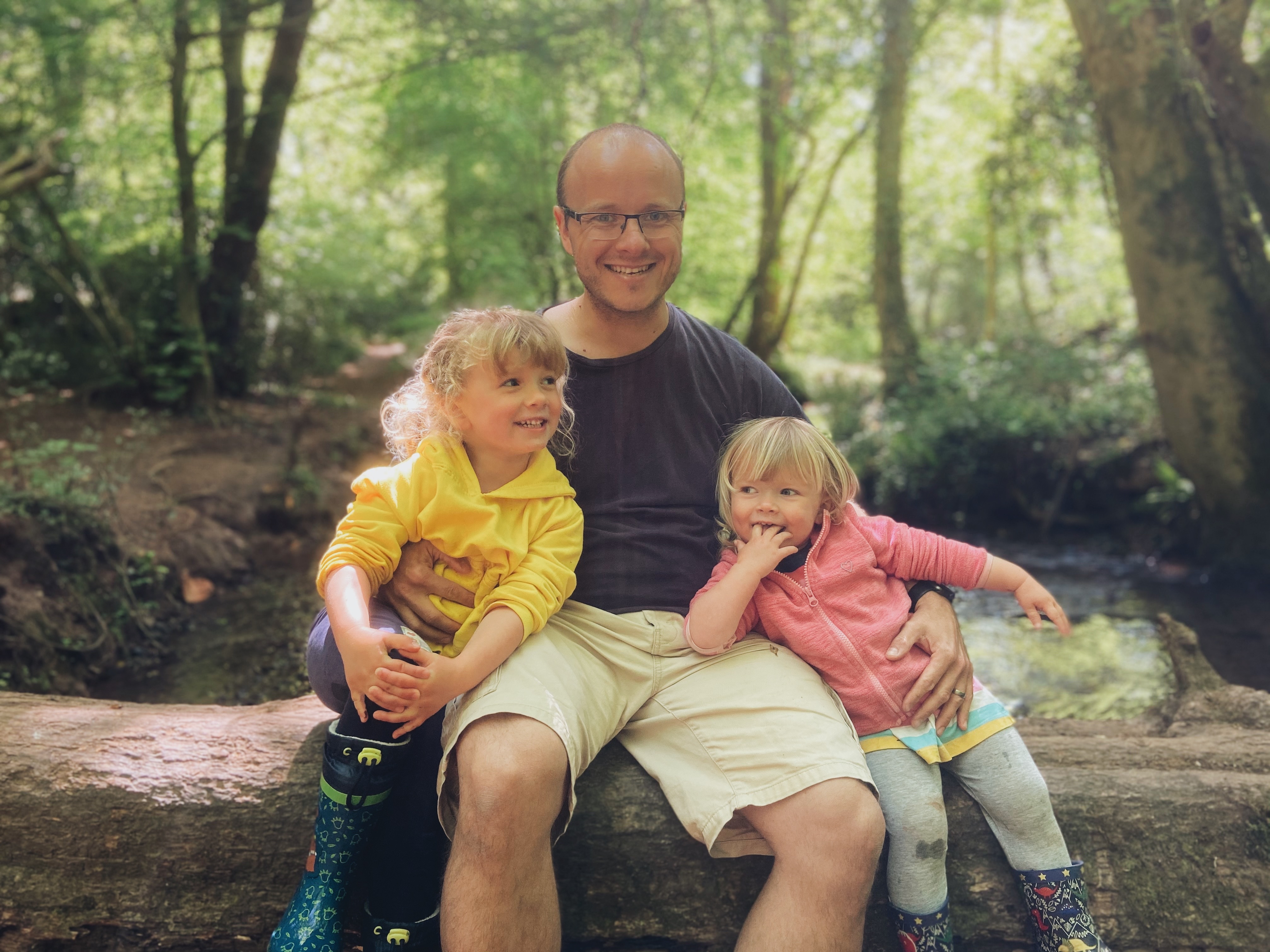 A dad and two girls sat on a log over a stream